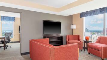 Habitación de hotel con televisor de pantalla plana montado en la pared, dos sillones tapizados en rojo, una mesita con lámpara y vistas a la ciudad a través de amplios ventanales.