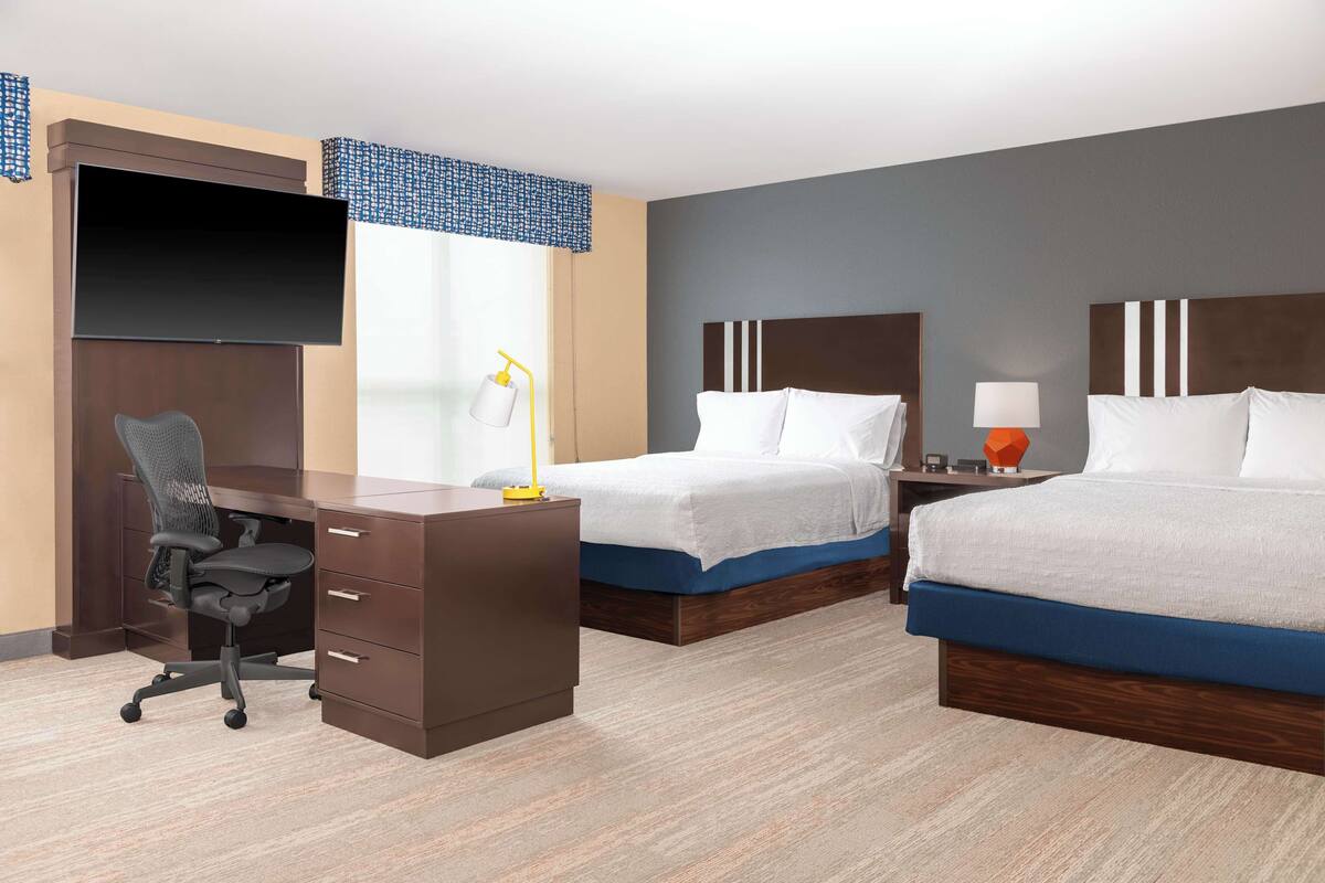 Habitación de hotel con escritorio, dos camas, televisor y ventana con vistas.
