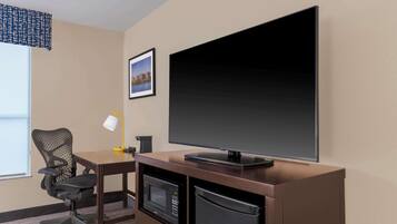 A hotel room with a desk, a flat-screen TV, a microwave, a refrigerator, a chair, and a window with a view of a cityscape.