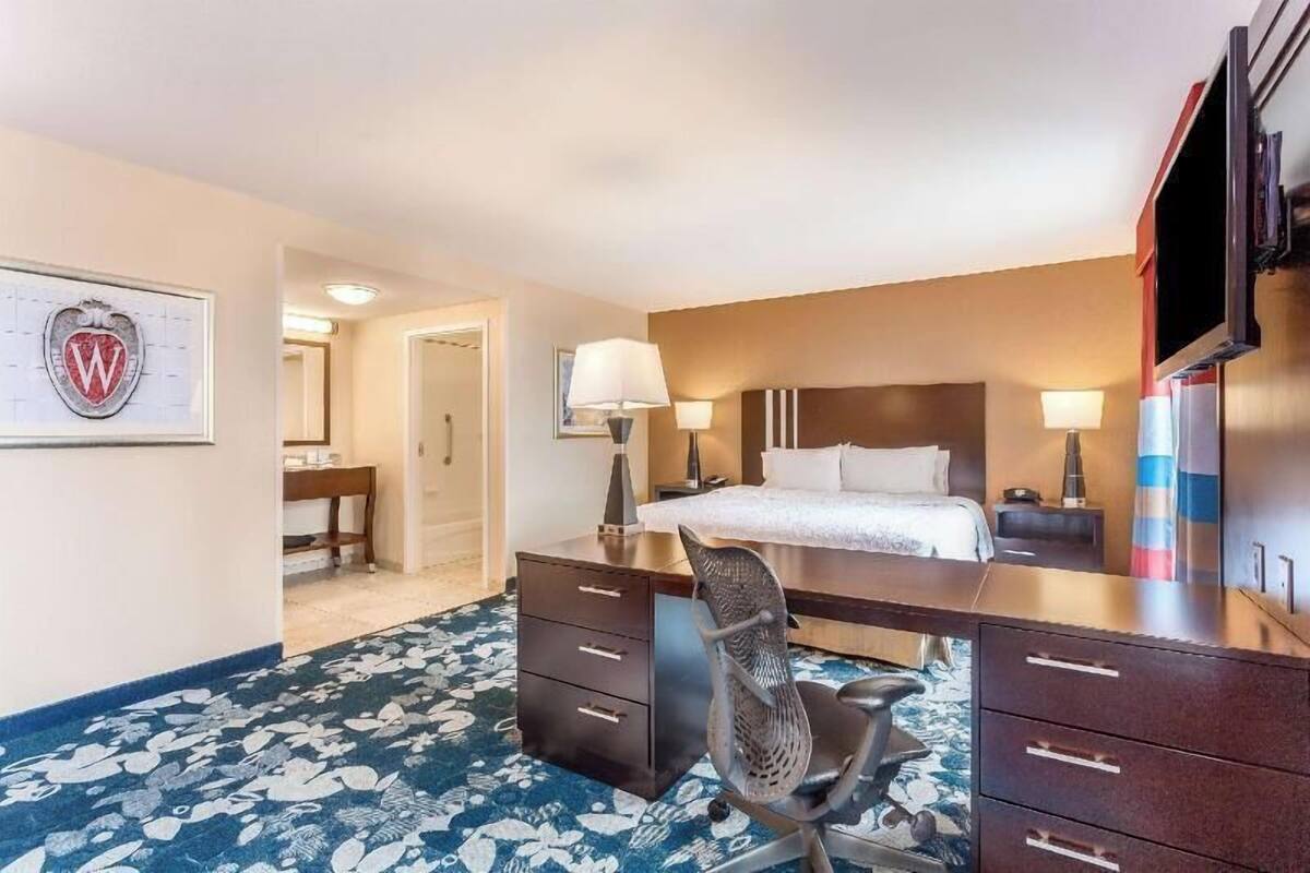 Quarto de hotel com cama, escrivaninha, cadeira e televisão.