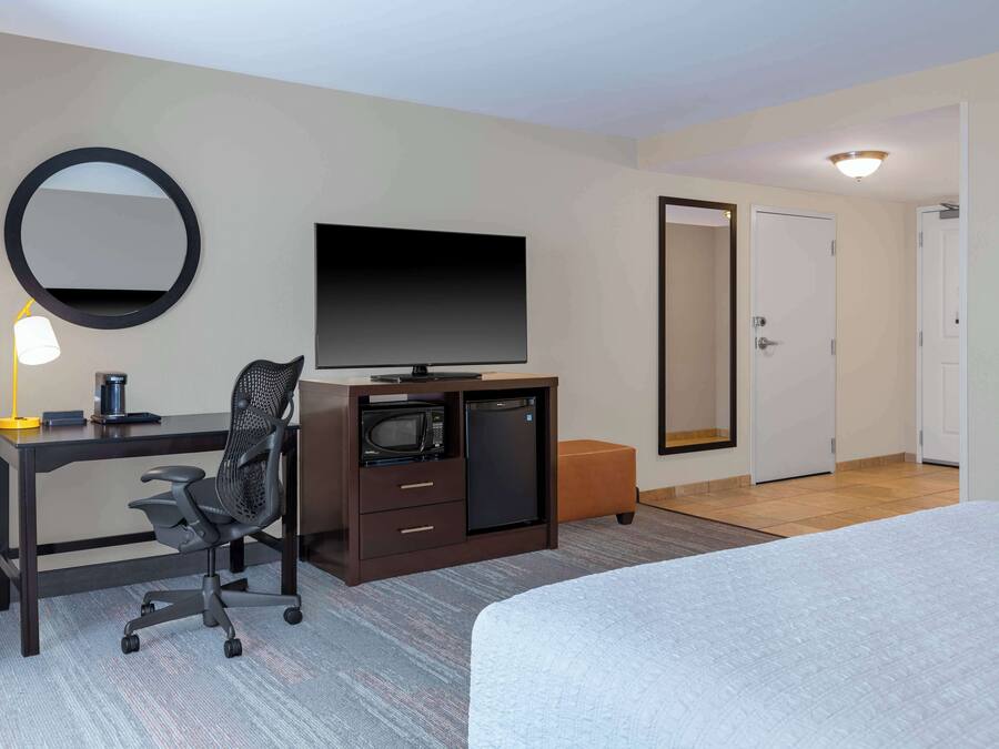 A hotel room with a bed, a desk with a computer, a television, a microwave, and a small refrigerator. There is a door leading to another room and a mirror on the wall.