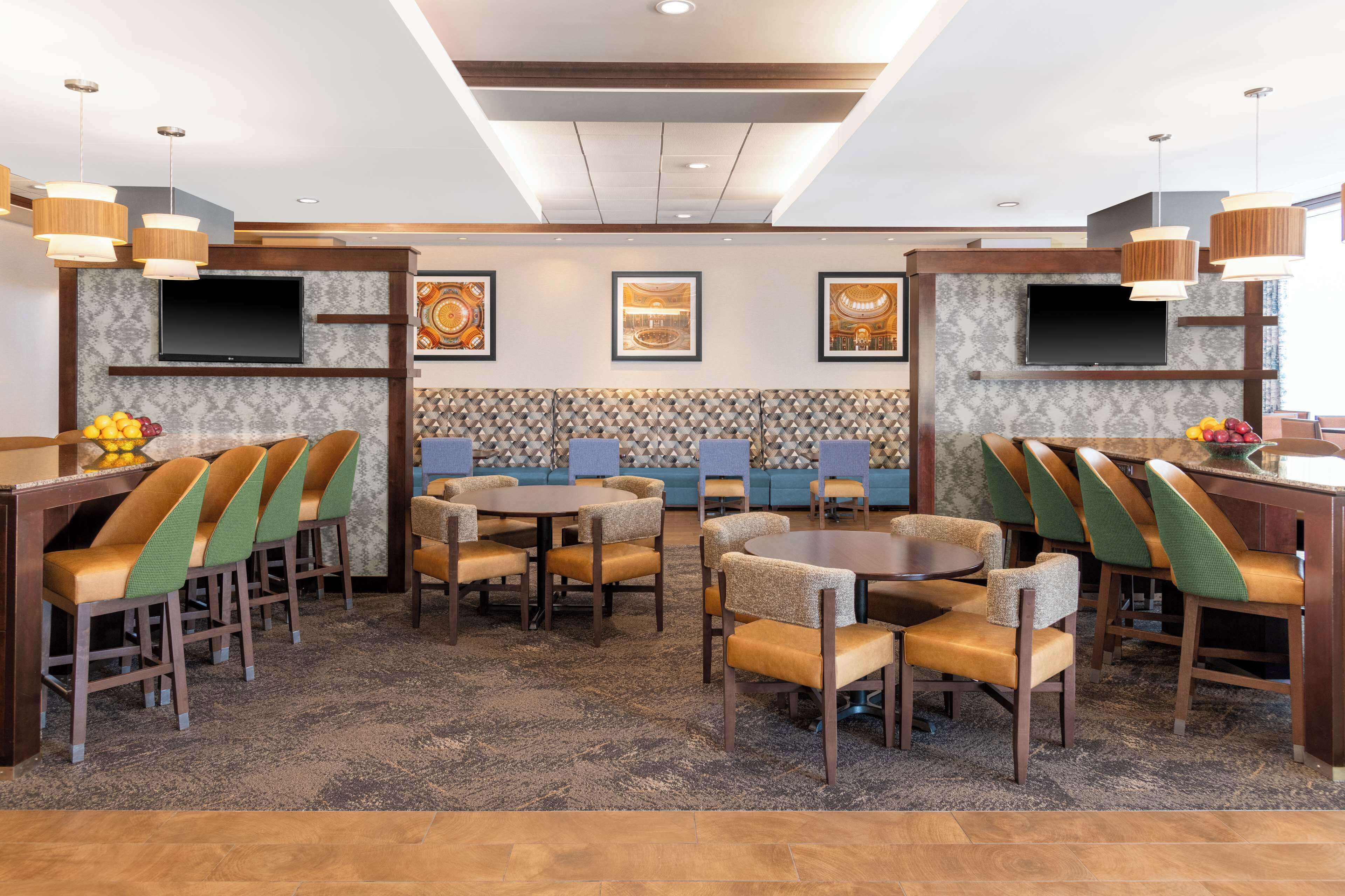 Un hall de hotel con varias zonas de estar, incluyendo mesas redondas y asientos de bar, con un televisor montado en la pared y obras de arte enmarcadas sobre los asientos.