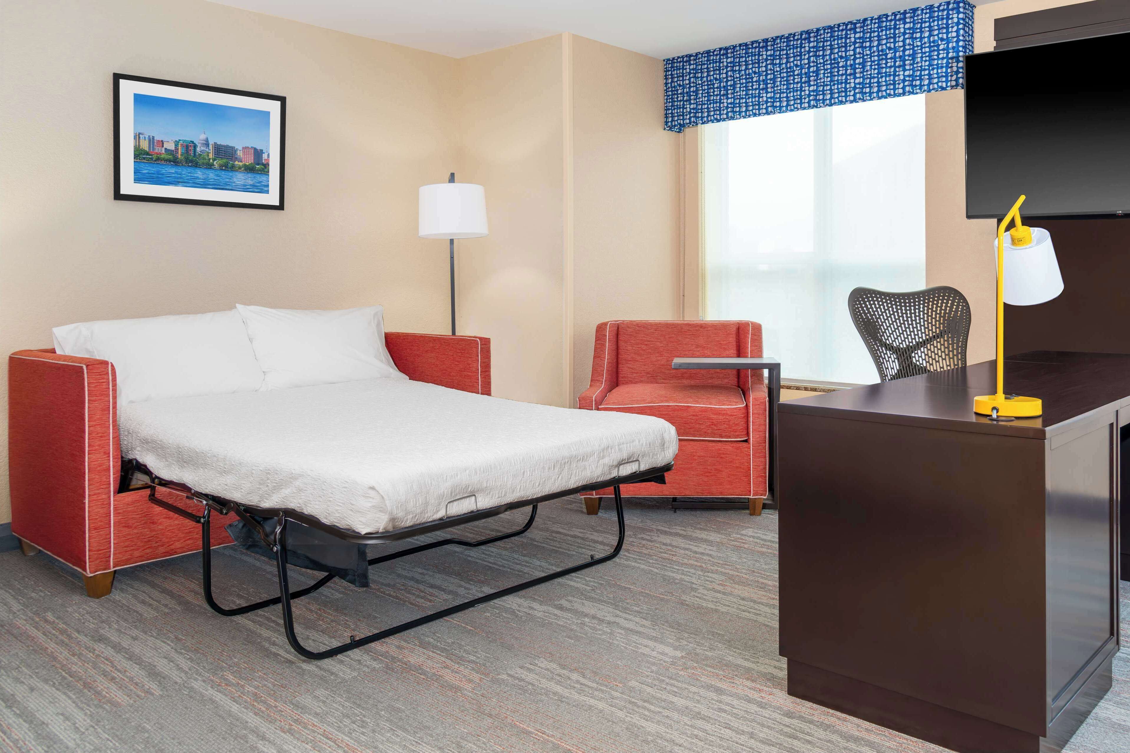 A hotel room with a bed, a desk, a chair, a lamp, and a large window with a view of a cityscape.