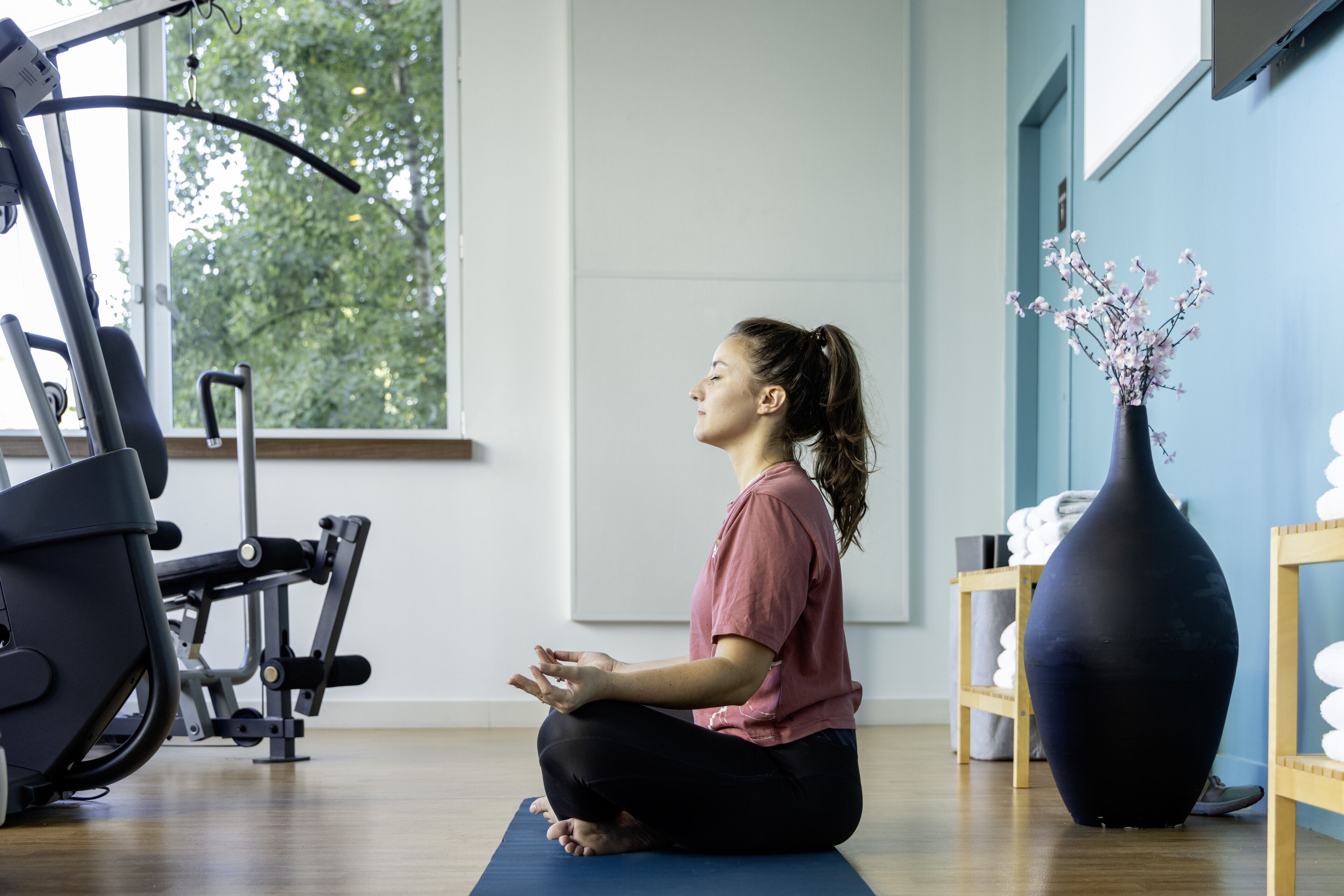 A person sitting on a yoga mat in a room with exercise equipment, a large vase with flowers, and a window with a view of trees.