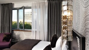 Hotelkamer met een bed, nachtkastje, lamp, raam met gordijnen en uitzicht op gebouwen.