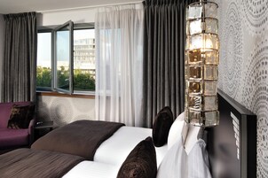 Habitación de hotel con cama, mesita de noche, lámpara, ventana con cortinas y vistas a edificios.