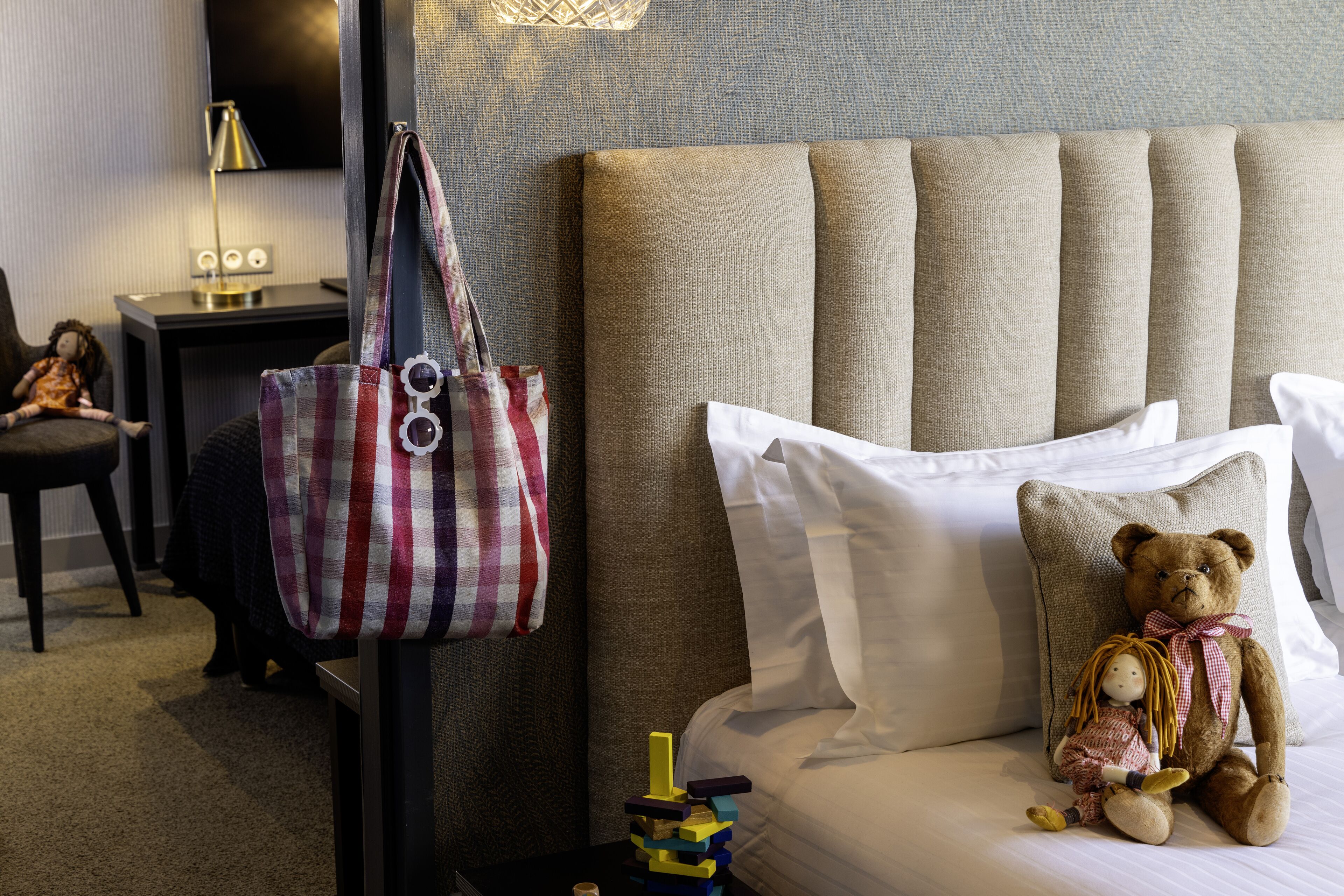 Una cama bien hecha con un osito de peluche y un bolso a rayas, una lámpara y una silla en una habitación de hotel.