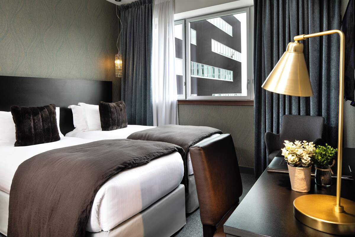 Habitación de hotel con una cama grande, un escritorio con una computadora portátil, una lámpara y vista a un edificio a través de la ventana.
