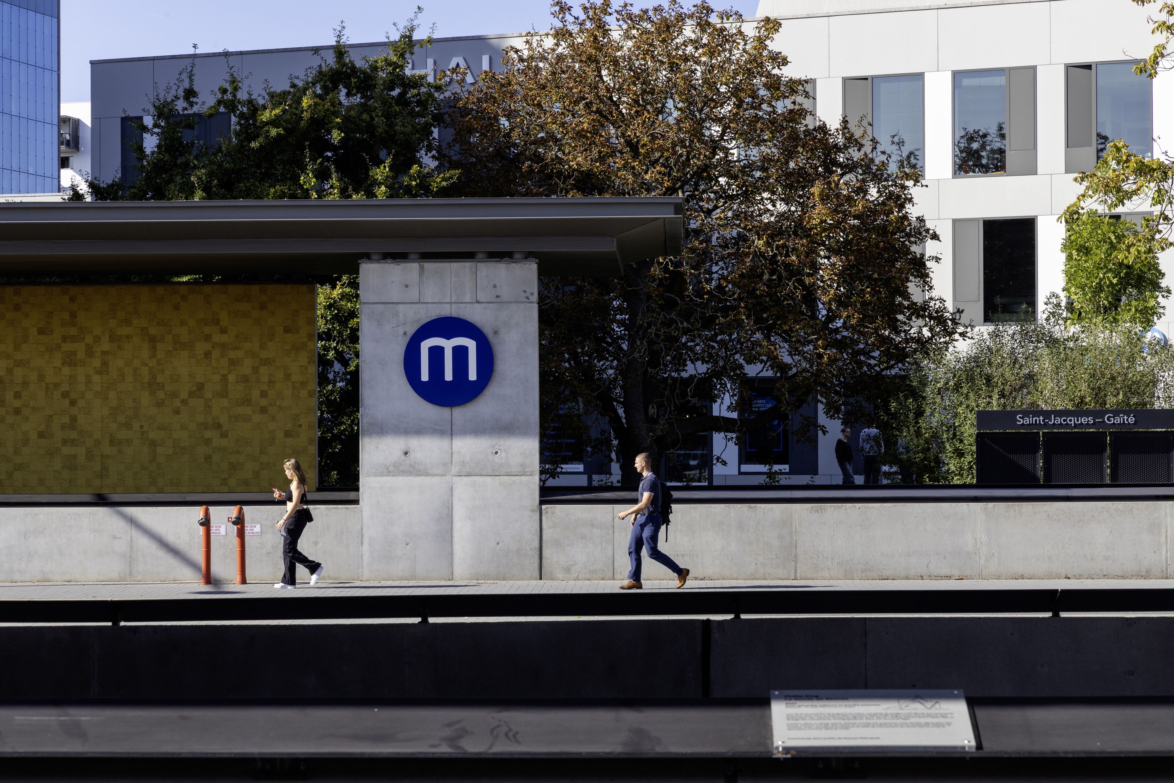 Um edifício moderno com um logotipo 'm' em destaque, uma passarela para pedestres e uma placa com o texto 'Saint-Jacques - Gaieté'.