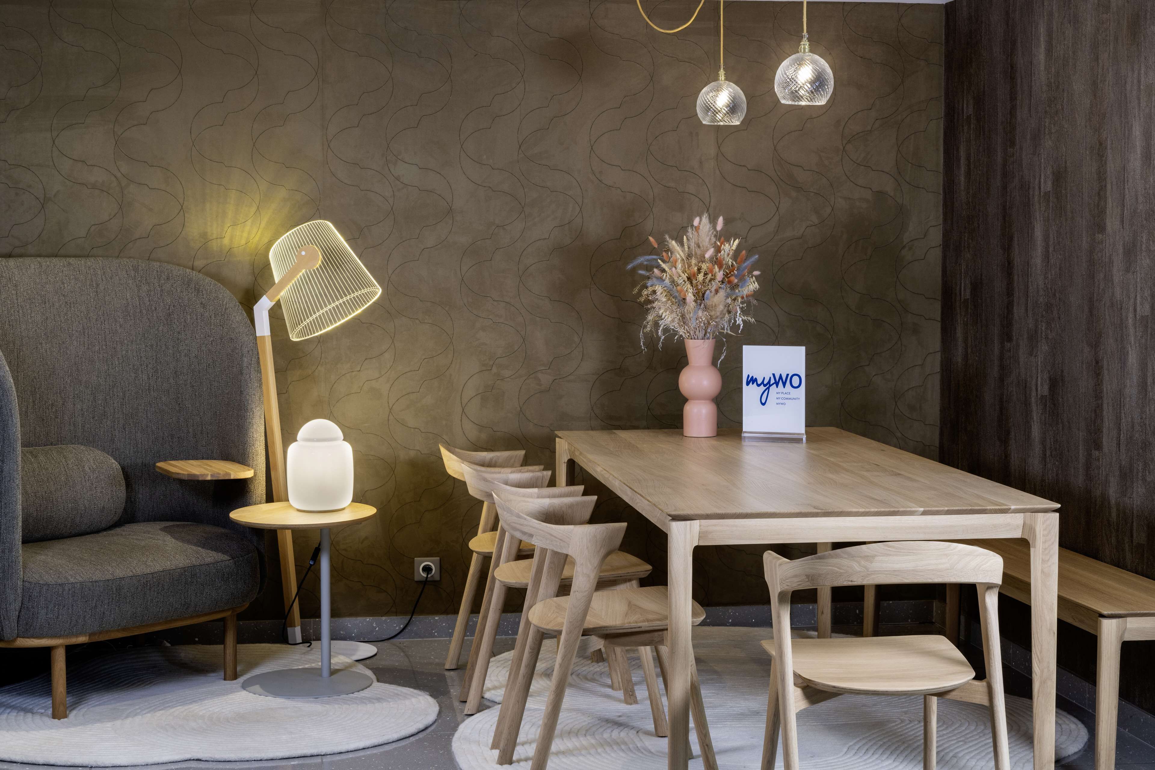 A modern room with a grey sofa, a wooden dining table with chairs, a small side table with a lamp, a vase with dried flowers, and a sign that reads "m2WO".