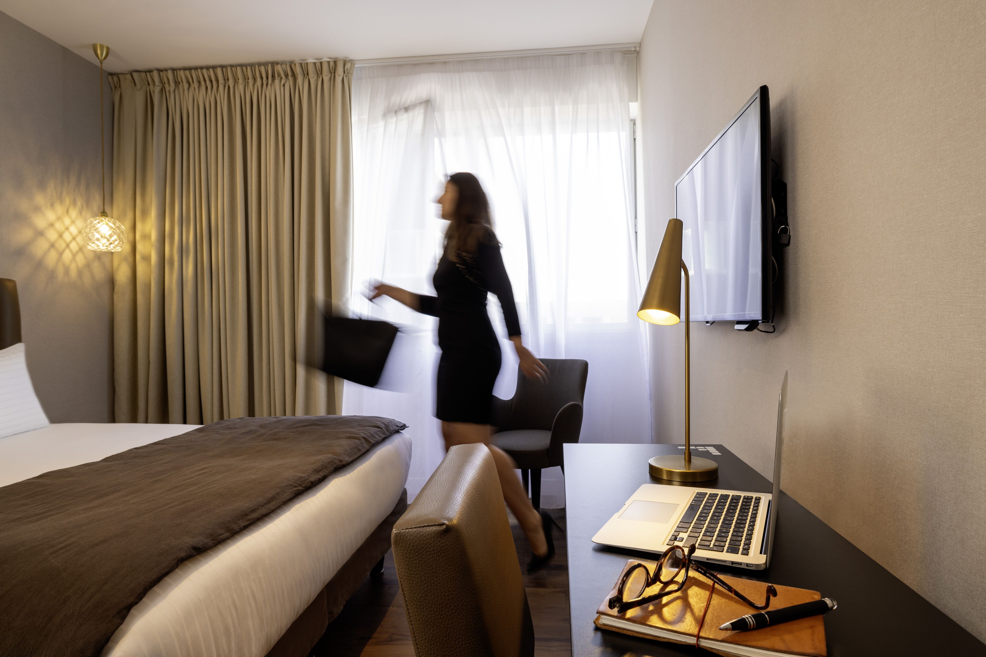 A hotel room with a bed, a desk with a laptop, a chair, and a TV on the wall.