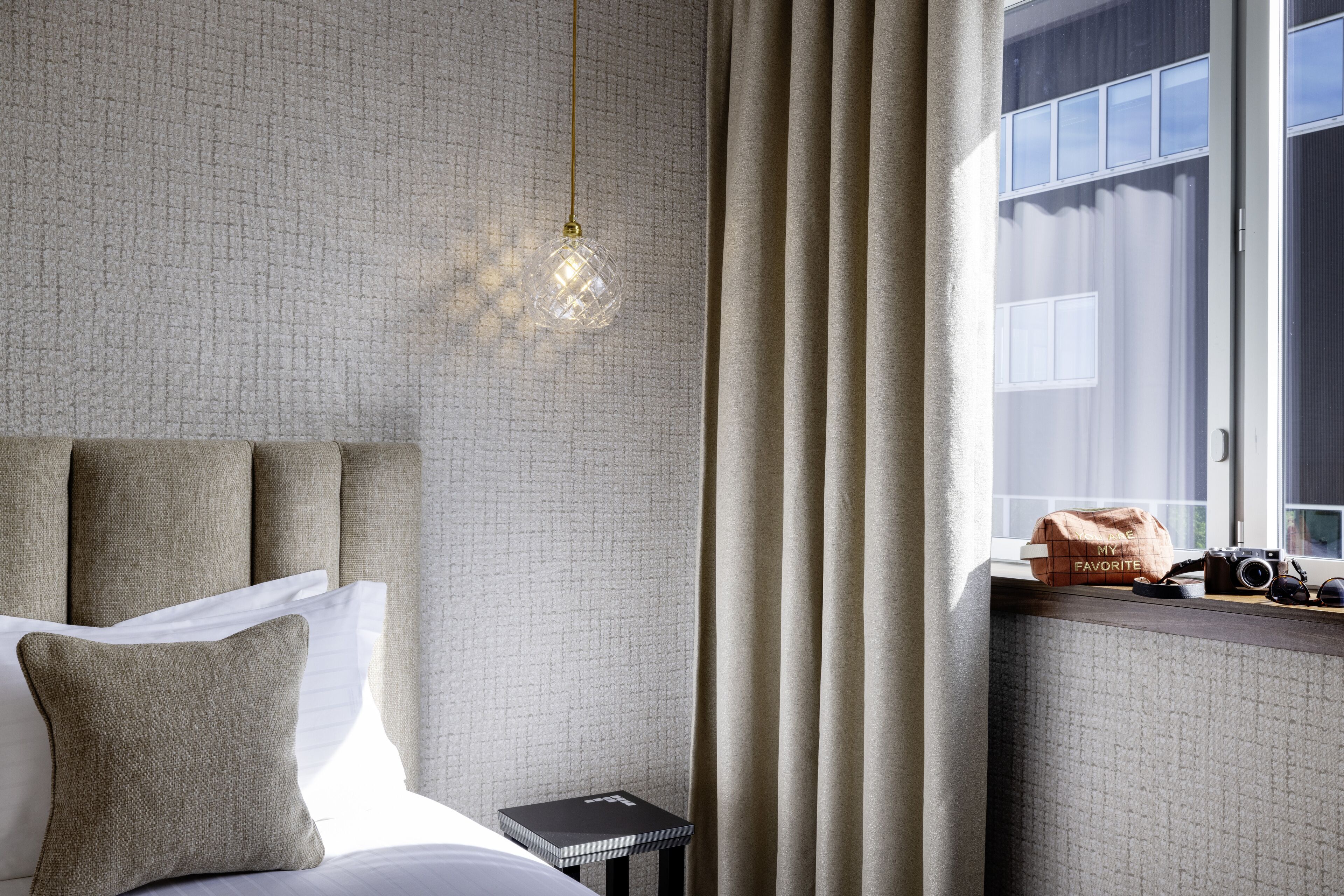 A neatly arranged hotel room with a bed, a bedside table with a book, a camera, and a bag labeled "MY FAVORITE" on the window sill.