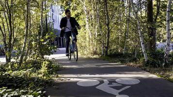 En cyklist på en asfalterad stig med en markering för cykelbana, omgiven av träd och en byggnad i bakgrunden.