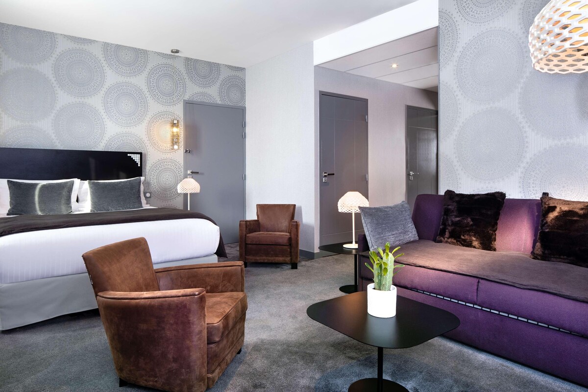 Una habitación de hotel moderna con una cama grande, un sofá, dos sillones, una mesita y una planta decorativa.