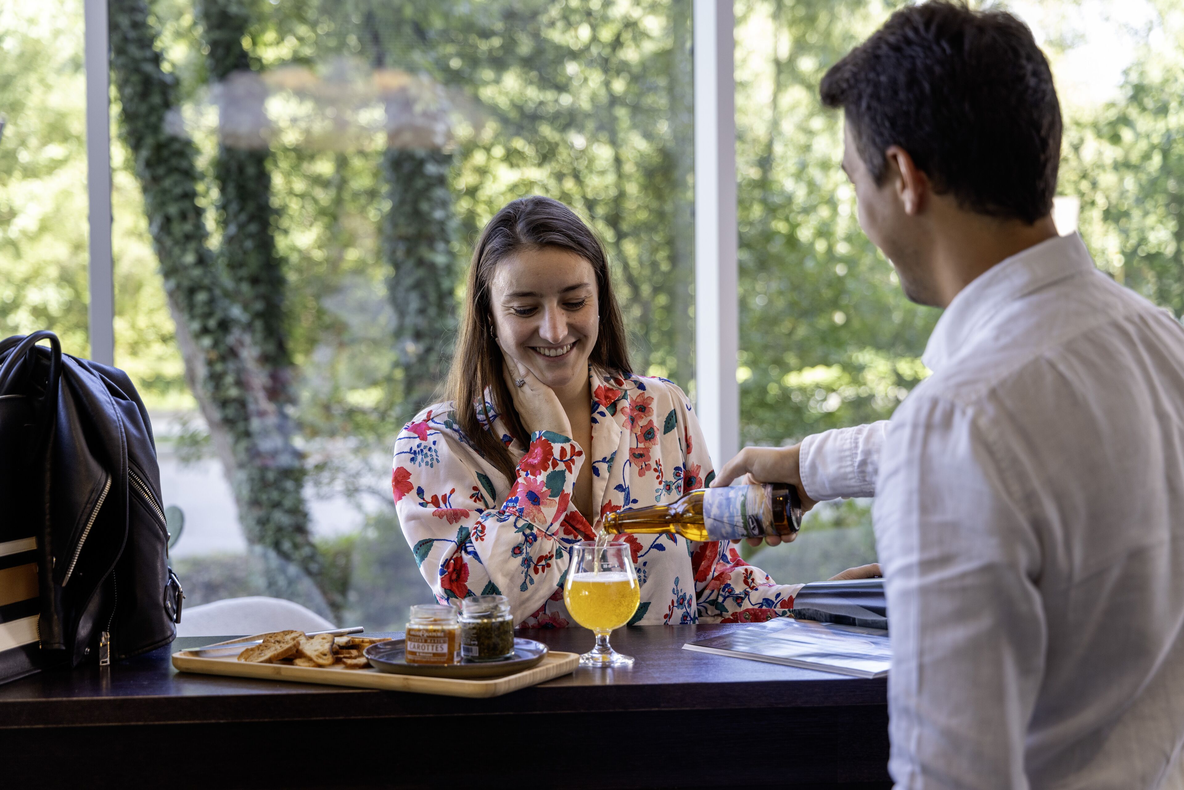 Una persona sirviendo una bebida en un vaso sobre una mesa con una computadora portátil, una bolsa y una bandeja con comida.