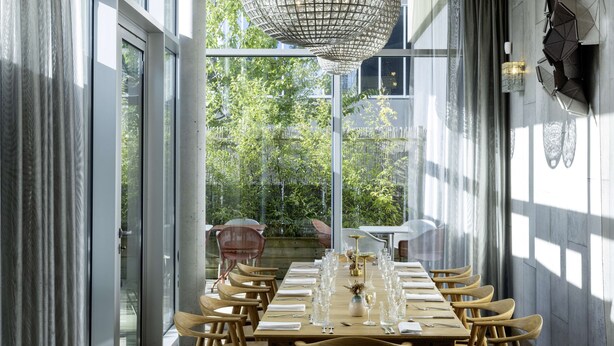 Een eetgedeelte met een lange tafel gedekt voor een maaltijd, grote ramen en een decoratieve hanglamp.