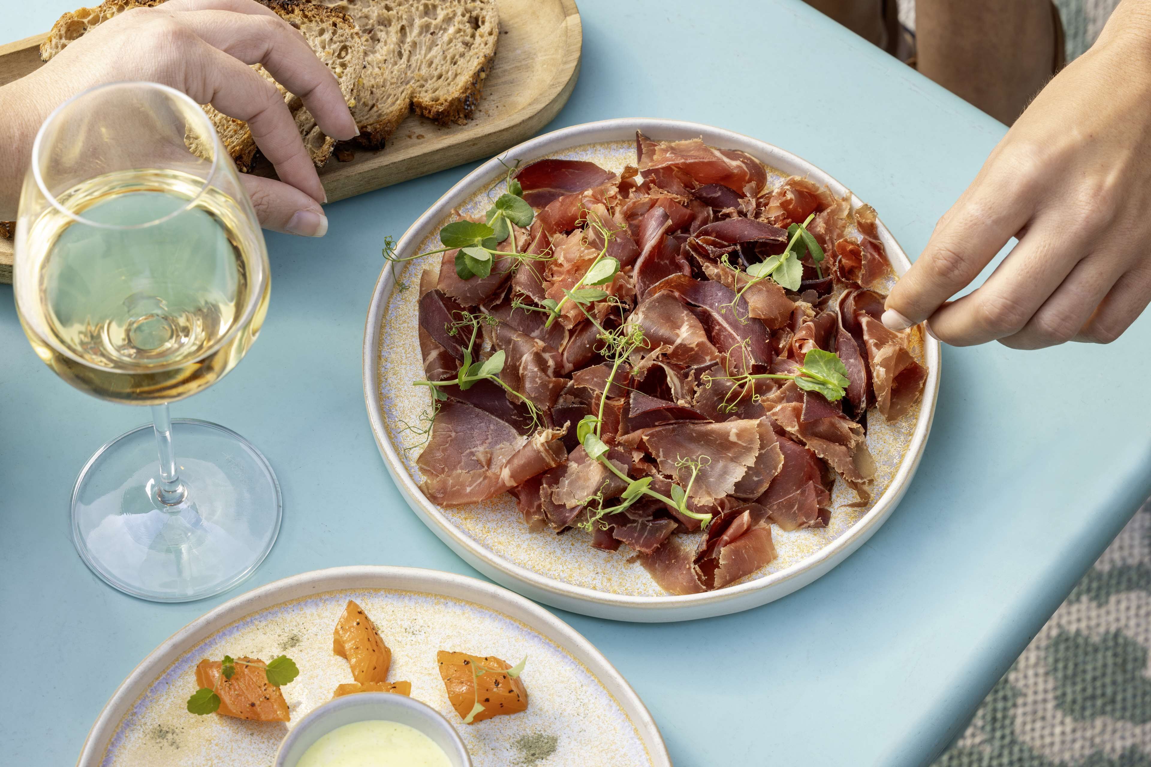 A dining table with a plate of prosciutto garnished with greens, a glass of white wine, and a bowl of soup.