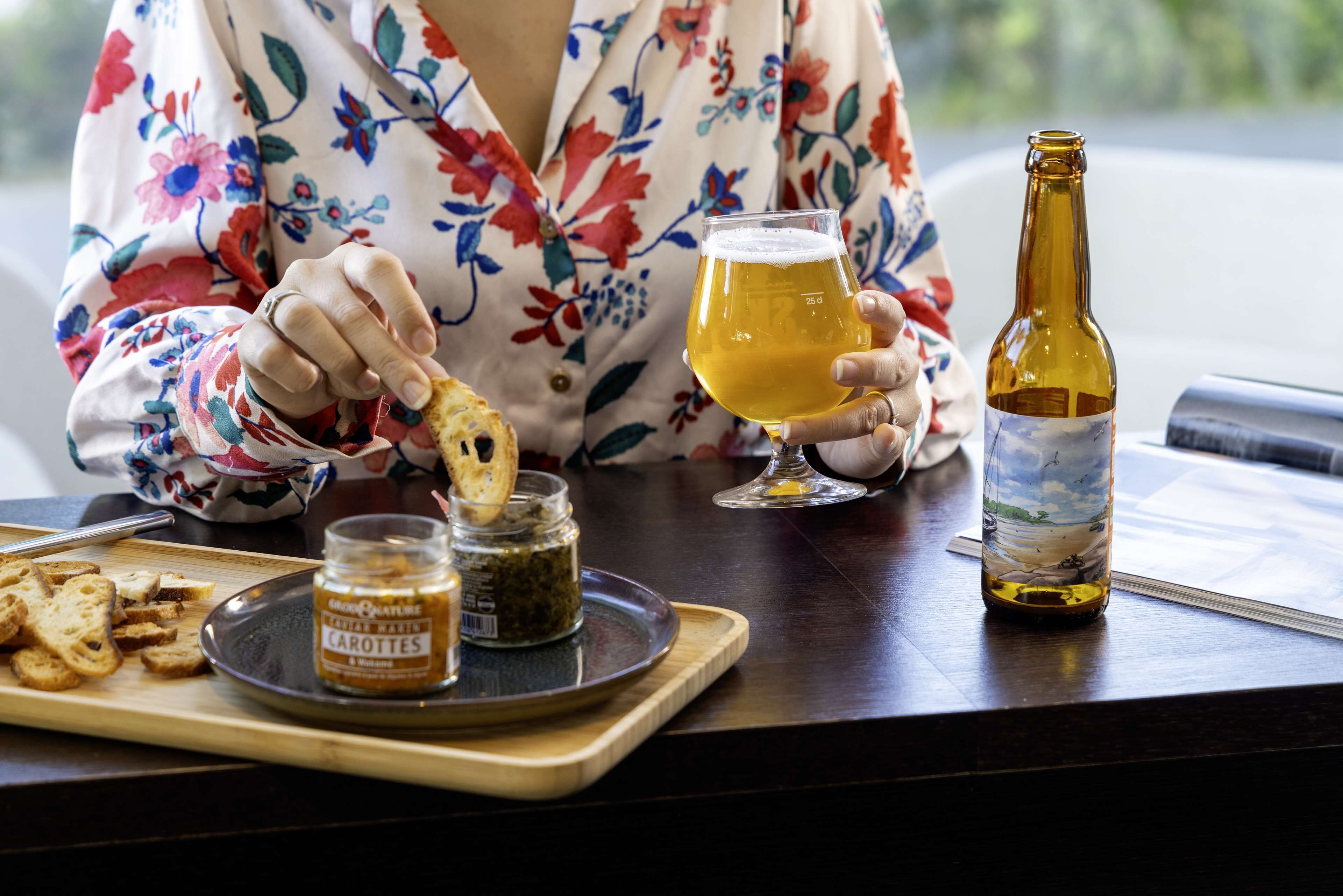 Una persona sosteniendo una jarra de cerveza y una botella sobre una mesa con una bandeja de comida y un frasco de mostaza.