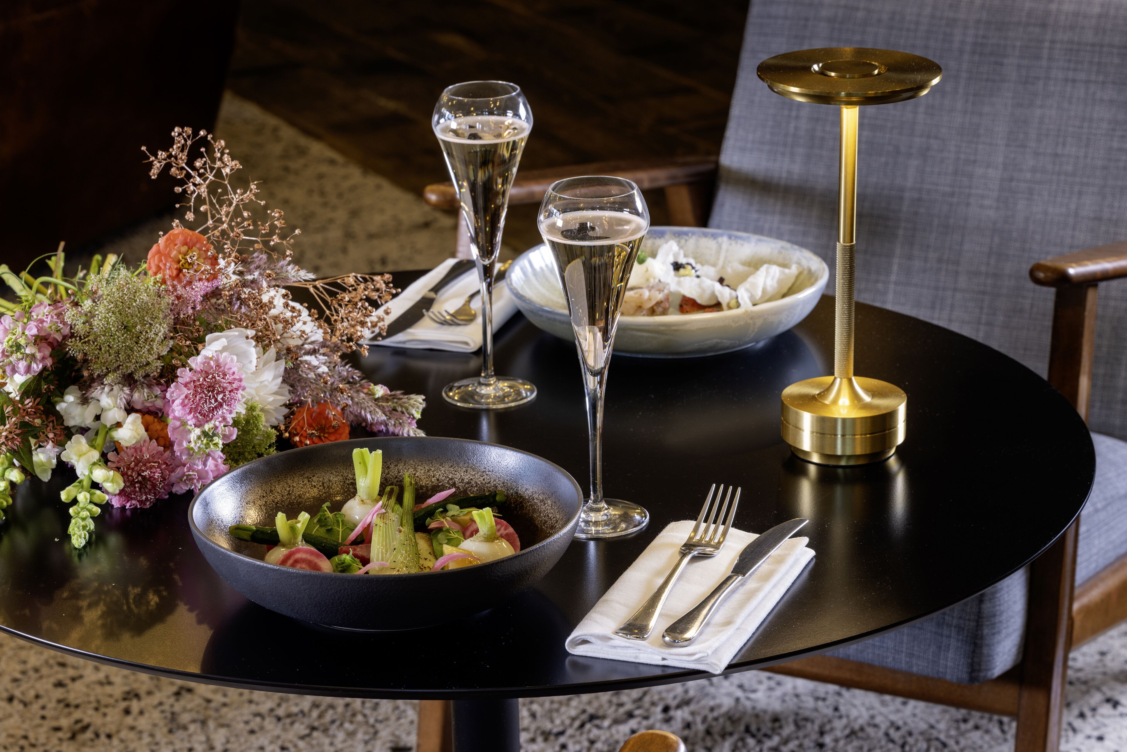 A dining table with a bowl of salad, two glasses of champagne, a plate of food, and a vase of flowers.