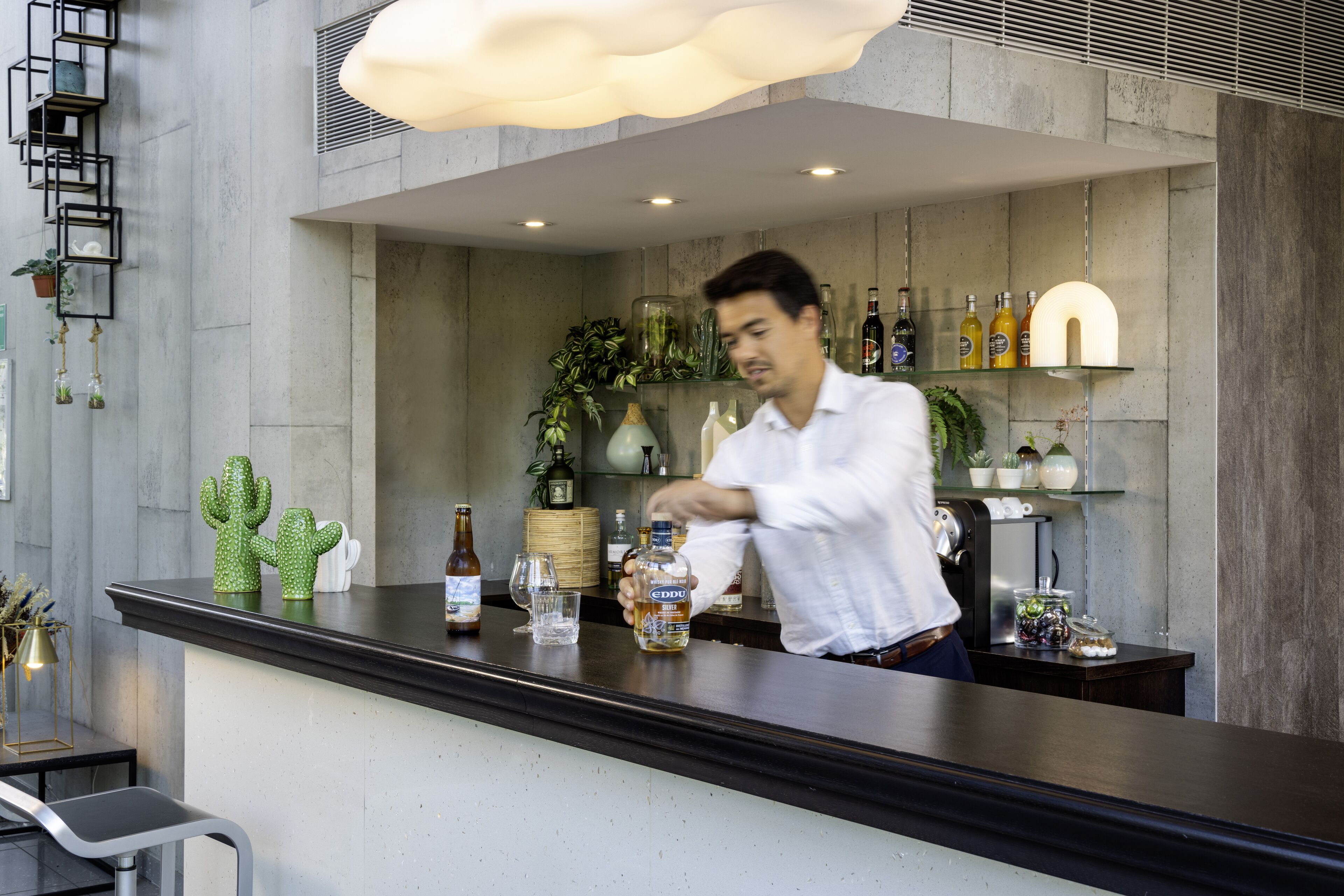 Zona de bar con un bartender preparando bebidas, estantes con botellas y plantas decorativas.