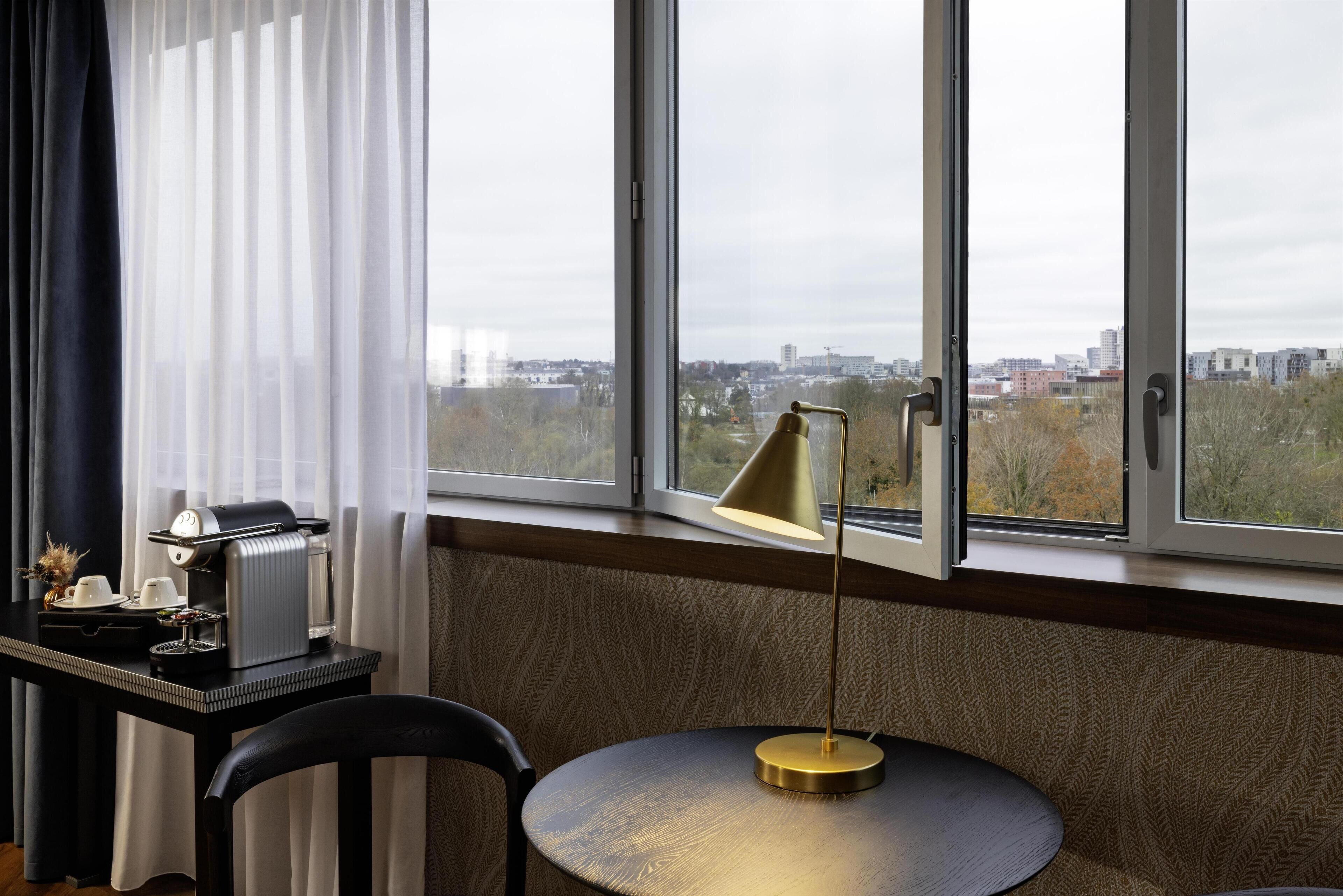 A room with a large window, a table with a coffee machine, a lamp, and a chair.