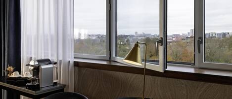 A room with a large window, a table with a coffee machine, a lamp, and a chair.