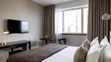 A hotel room with a bed, a desk, a chair, a TV, a small table with a lamp, and a window with curtains.