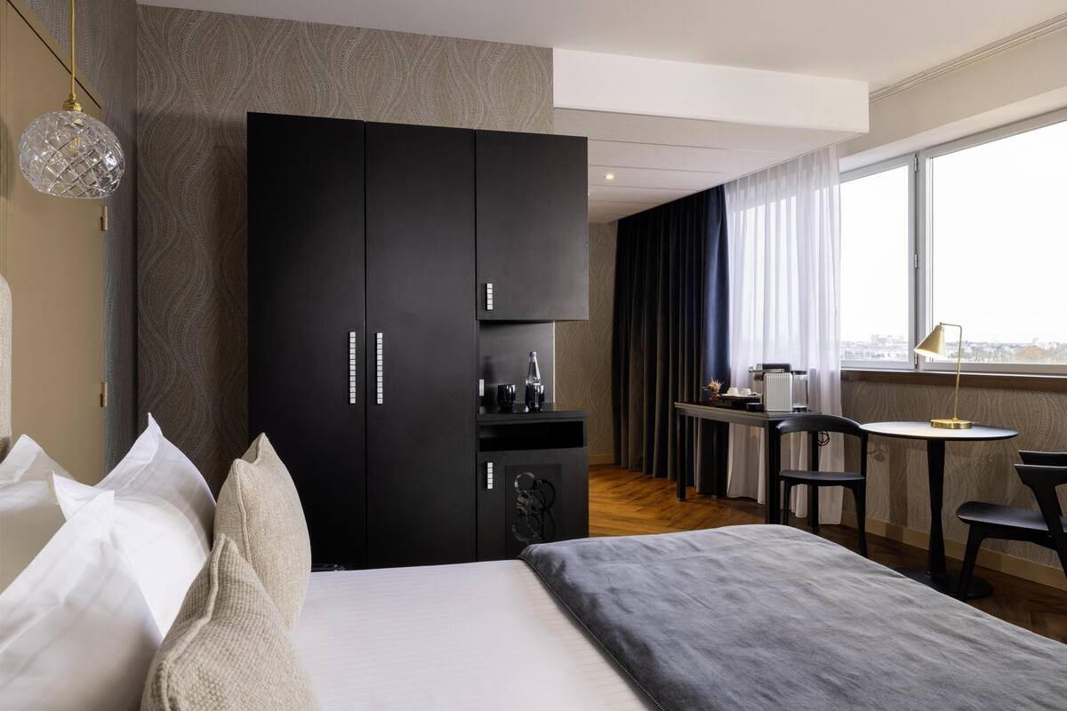 Een moderne hotelkamer met een groot bed, een kast, een kleine kitchenette, een zithoek met een tafel en stoelen, en een groot raam met uitzicht op de stad.
