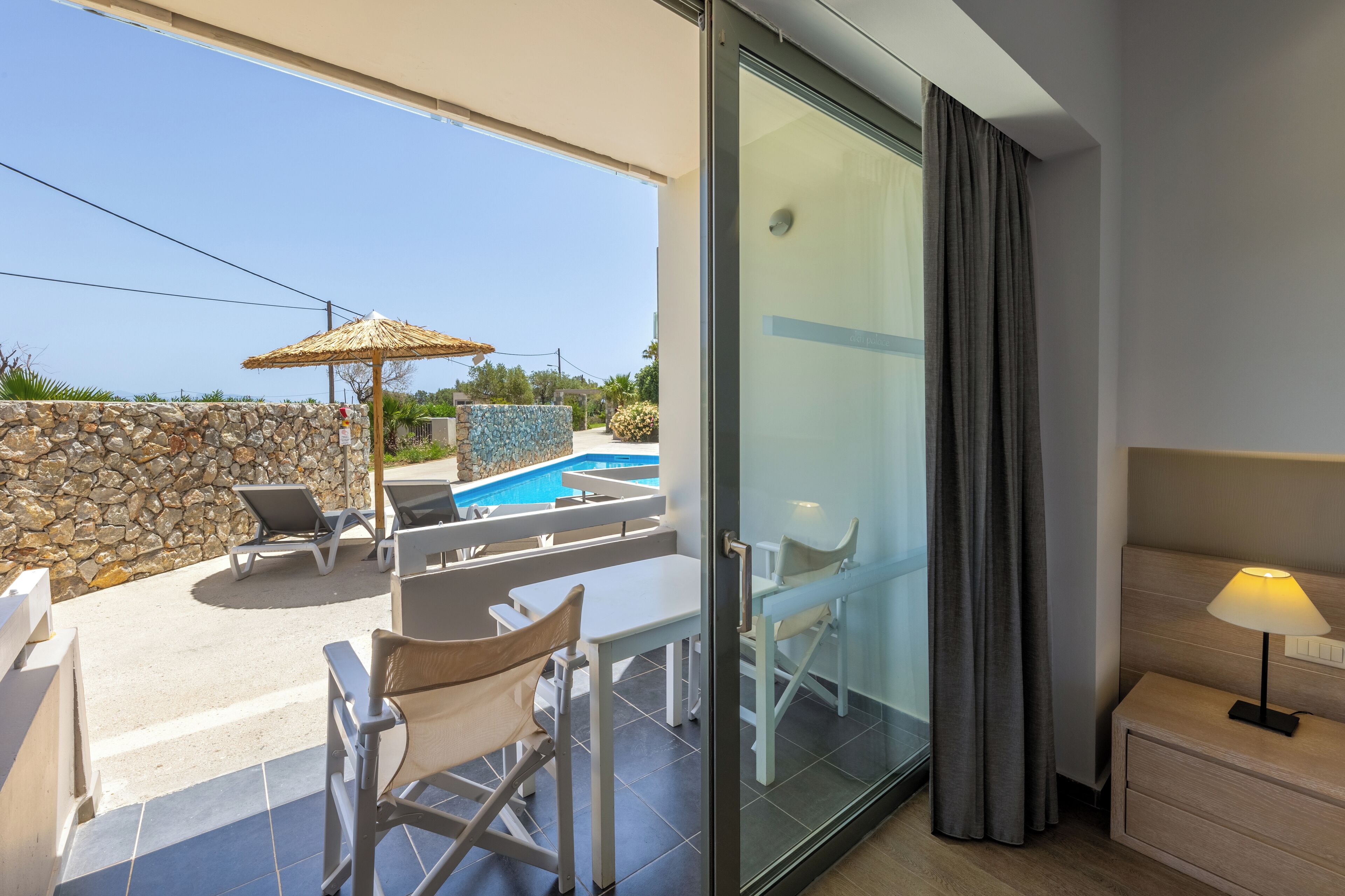 Habitación de hotel moderna con puerta de vidrio que da a un balcón con piscina, mobiliario exterior y pared de piedra.