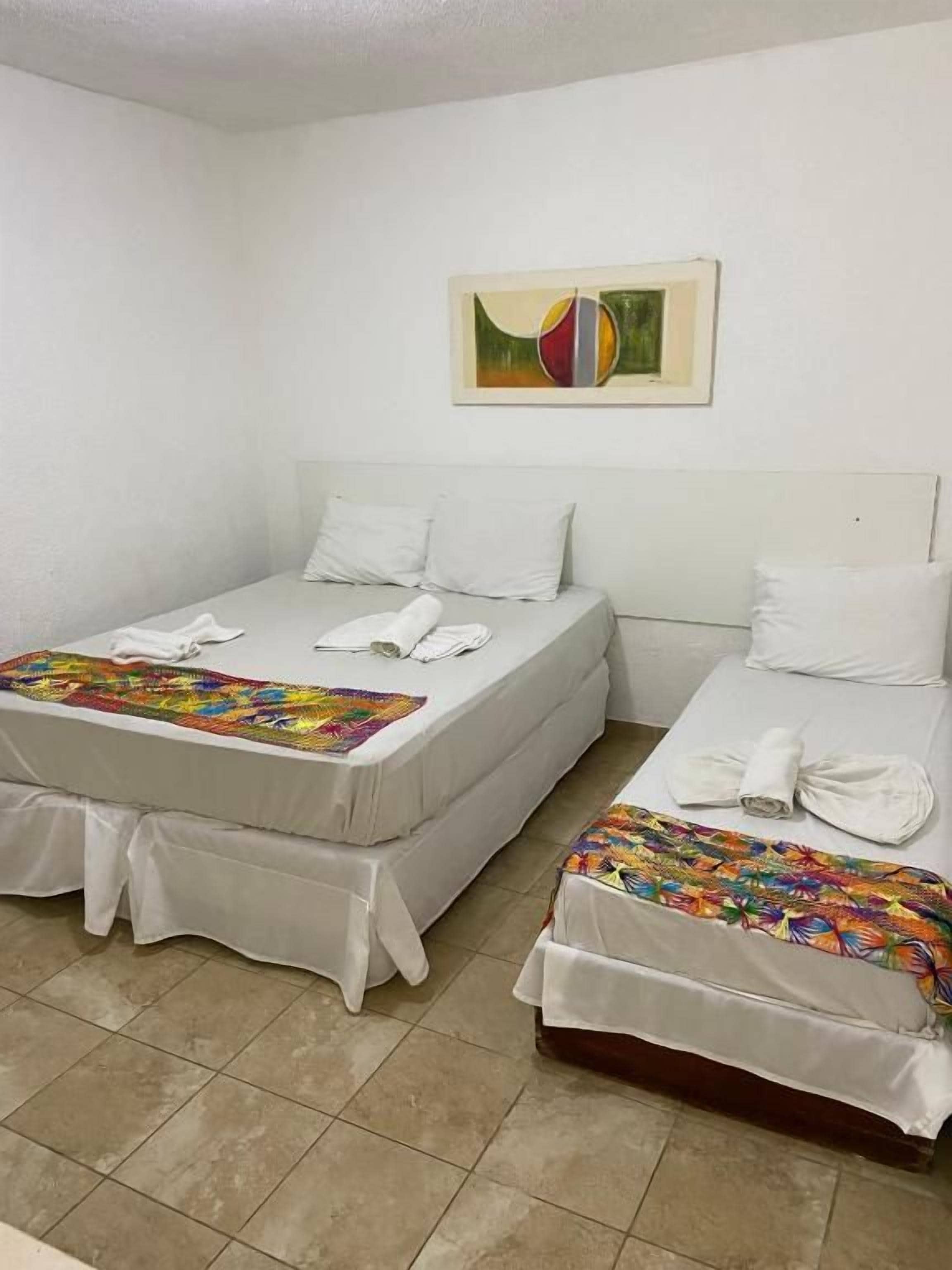Habitación de hotel con dos camas, cada una con cubrecama y almohadas blancas, y una colcha de colores sobre una de ellas.