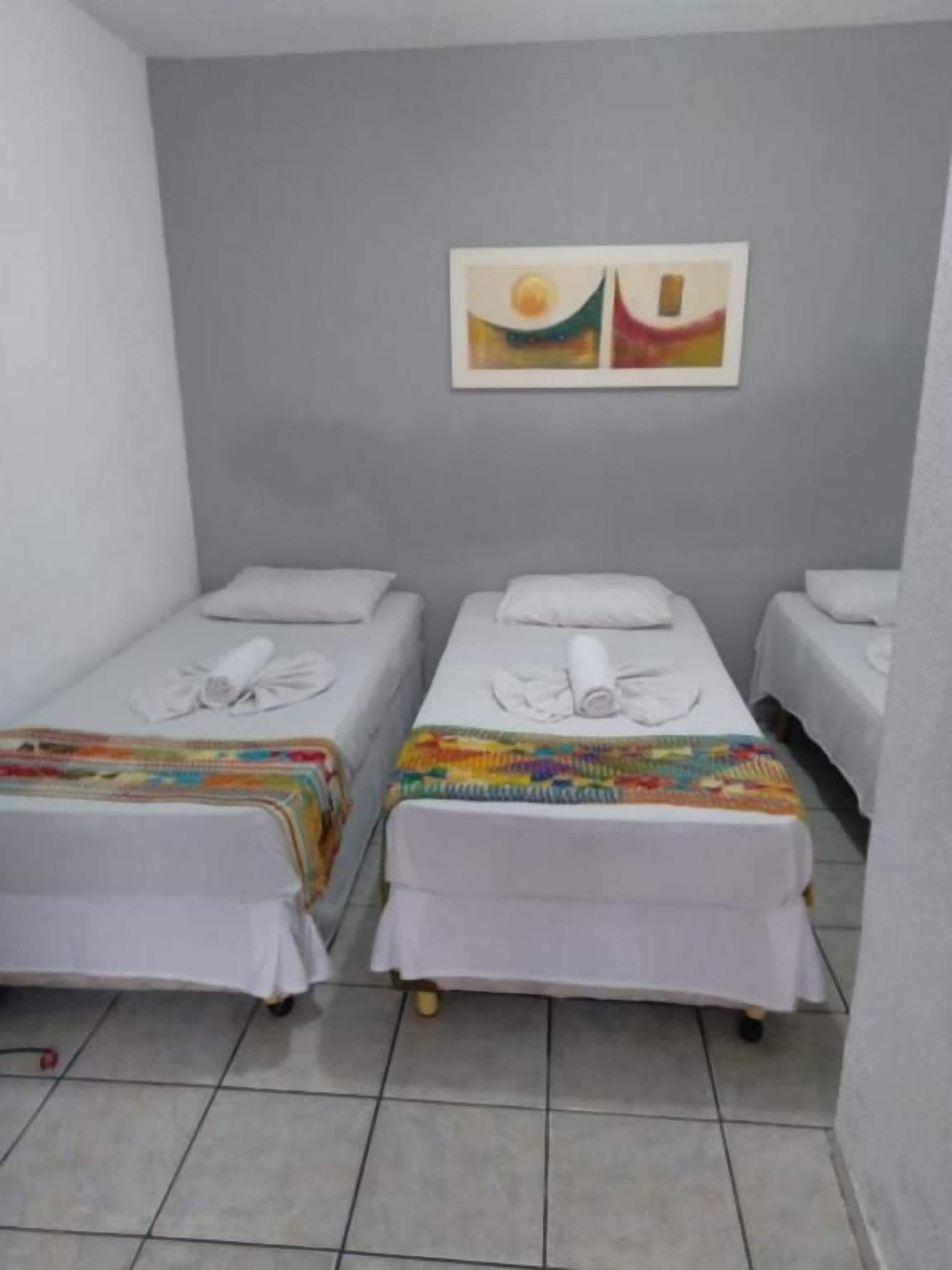 A room with three single beds, each with a white mattress and pillow, and a colorful blanket. There is a painting on the wall above the beds.