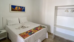 A hotel room with a bed, a nightstand, a painting on the wall, and a clothing rack. - Pousada Nossa Casa (Maceió)