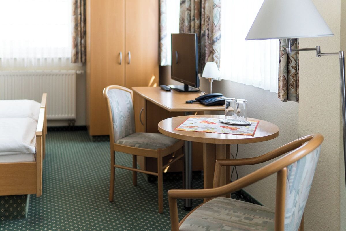 A hotel room with a bed, a desk with a computer, a chair, and a table with a lamp.