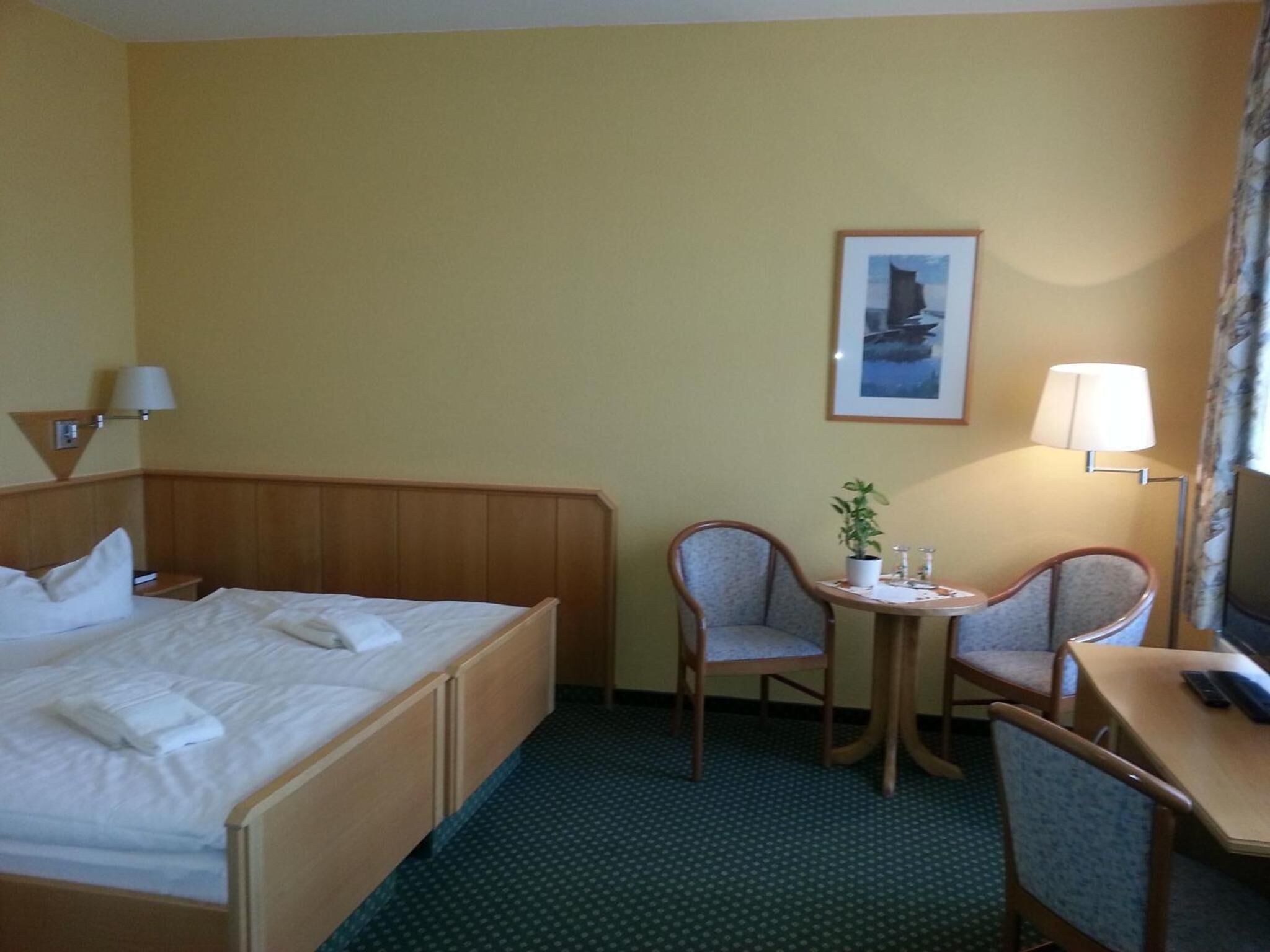 Habitación de hotel con cama, dos sillas, una mesita con una planta, lámpara y un cuadro en la pared.