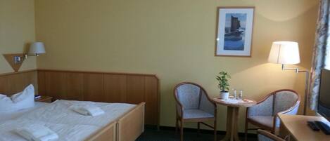 Habitación de hotel con cama, dos sillas, una mesita con una planta, lámpara y un cuadro en la pared.