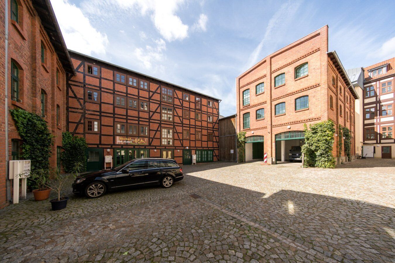 A courtyard with cobblestone pavement, a black car parked in the middle, and a building with a sign that reads "Hotel".