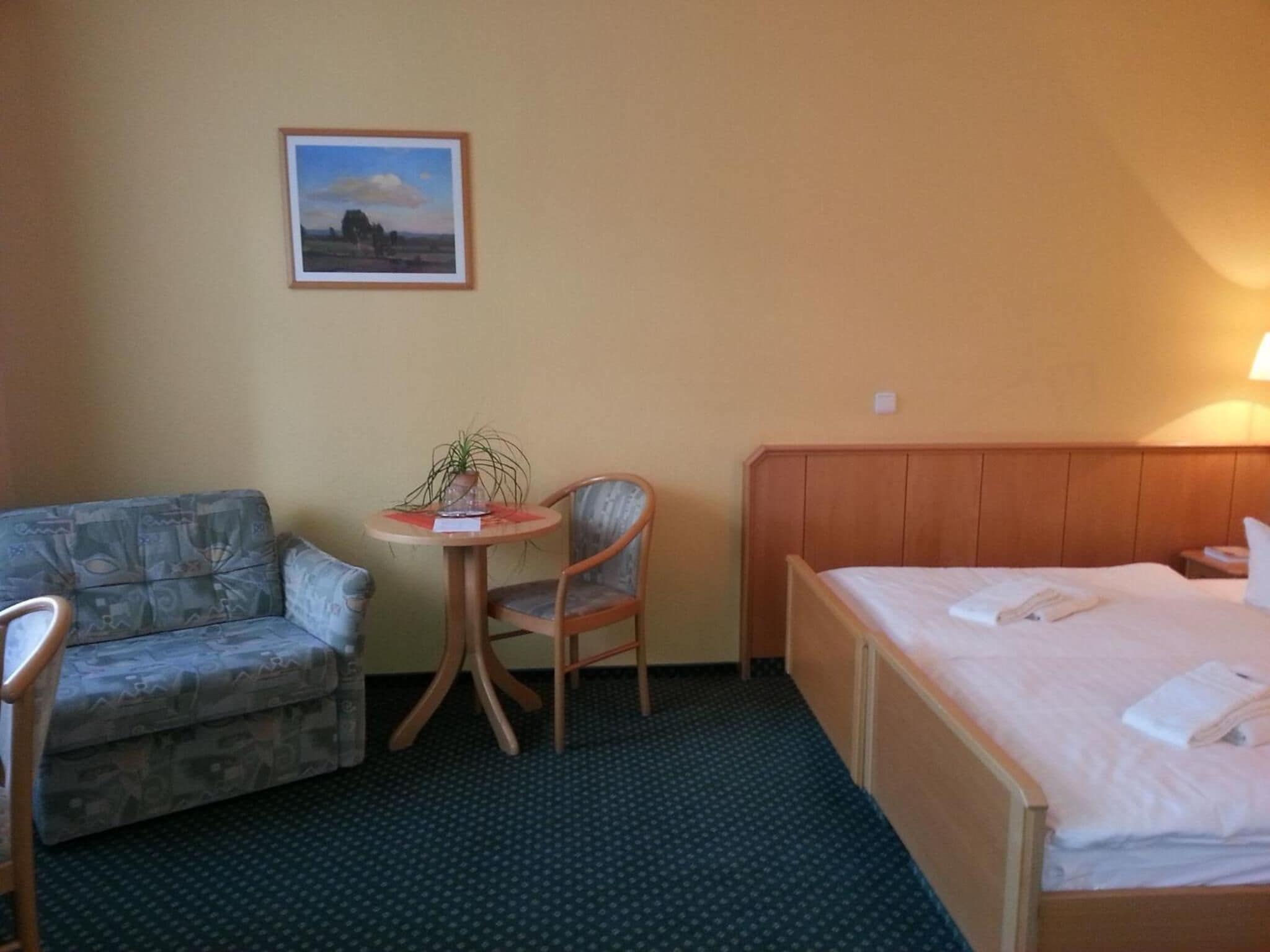 Et hotelværelse med en seng, en stol, et lille bord med en plante og et billede i ramme på væggen.