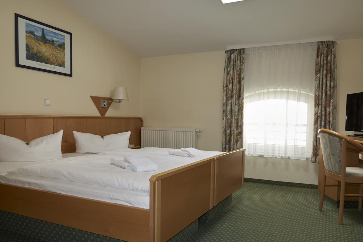A hotel room with a wooden bed, a desk, a chair, a window with curtains, a radiator, and a framed painting on the wall.