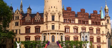 Un edificio histórico con una torre destacada, arquitectura ornamentada y un jardín cuidado con una fuente central.
