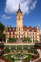 Un edificio histórico con una torre destacada, arquitectura ornamentada y un jardín cuidado con una fuente central.