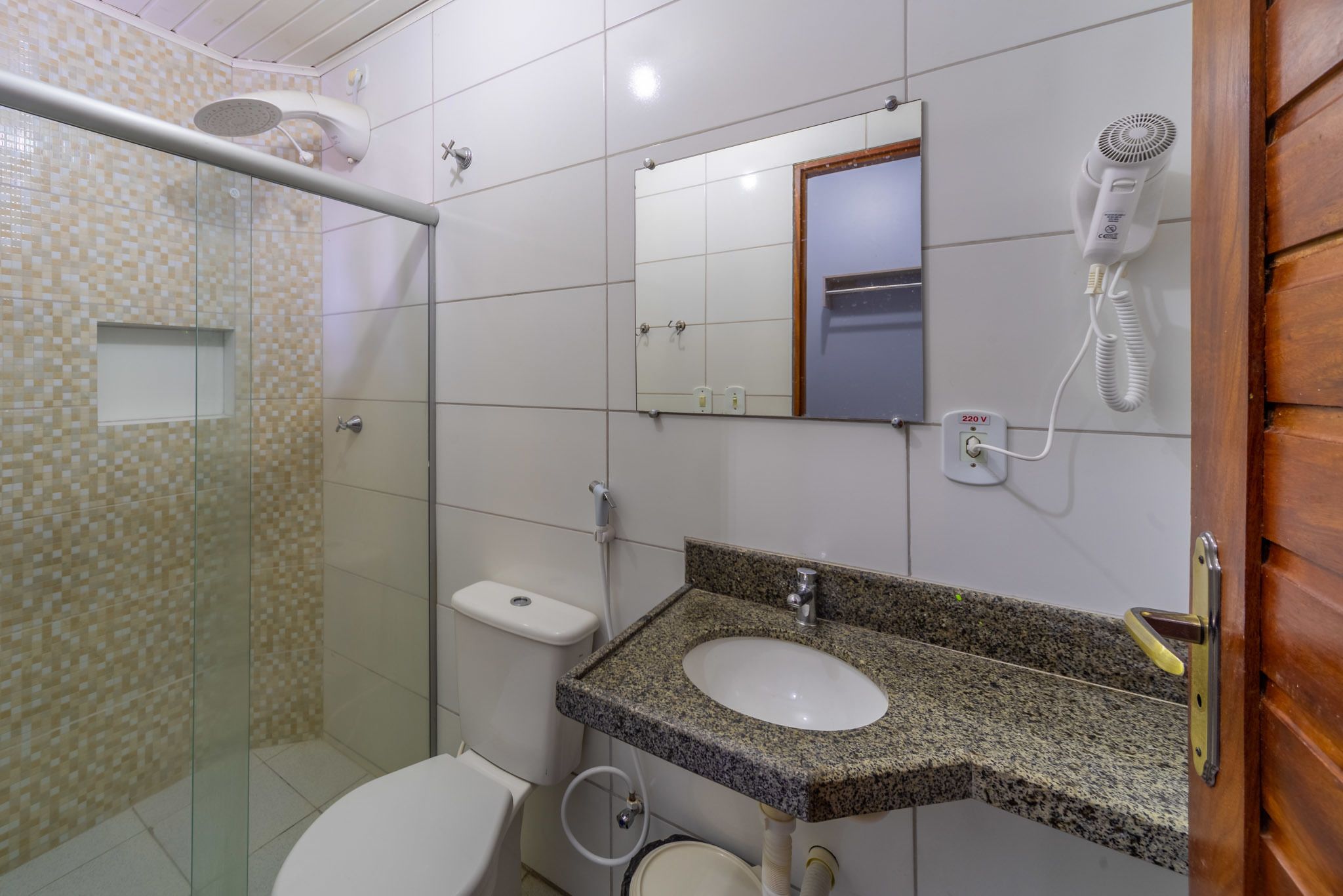 A bathroom with a glass shower enclosure, a white toilet, a granite countertop sink, a mirror, and a hairdryer mounted on the wall.