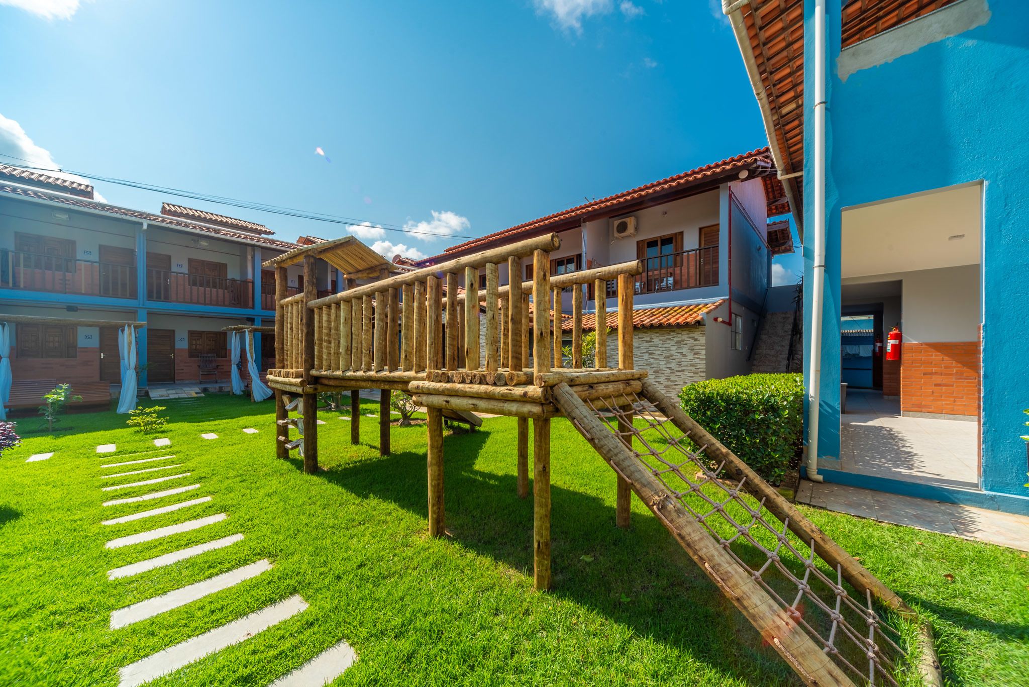 A playground structure with a wooden slide and climbing net, surrounded by a well-maintained lawn and a building with a red-tiled roof.