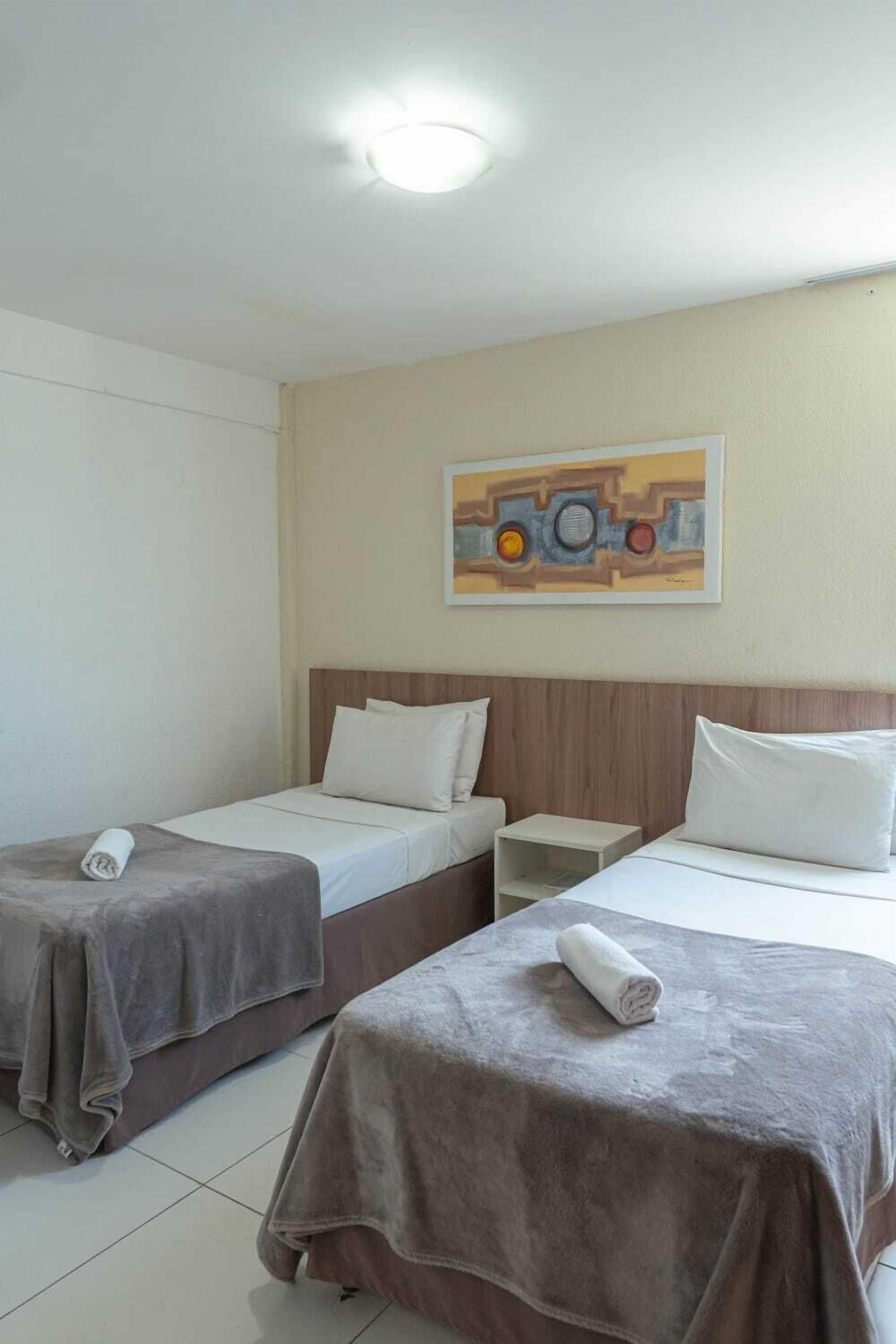 A hotel room with two beds, each with a brown bedspread and white pillows, a small bedside table, and a wall-mounted artwork.