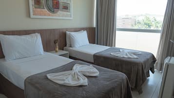 A hotel room with two beds, a nightstand, a painting on the wall, and a view of the city through the window.