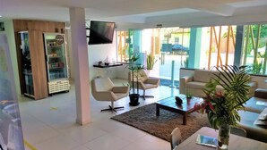 Ein modernes Wohnzimmer mit Flachbildfernseher, zwei Sofas, einem Couchtisch und einem Esstisch. Ein Kühlschrank mit Glastür und ein Regal mit verschiedenen Gegenständen sind ebenfalls vorhanden.