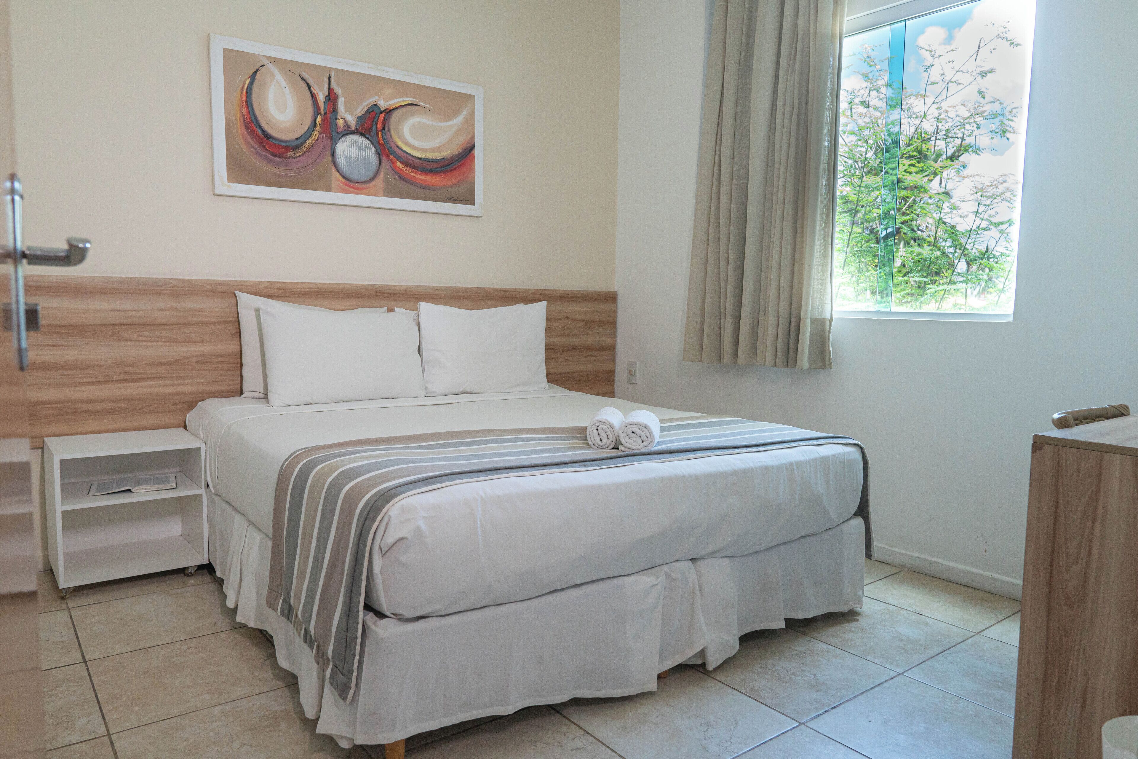 A neatly made bed with white linens and a striped blanket, a bedside table with a lamp, a painting on the wall, and a window with curtains.