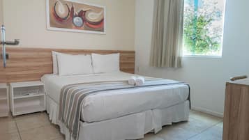 A neatly made bed with white linens and a striped blanket, a bedside table with a lamp, a painting on the wall, and a window with curtains.