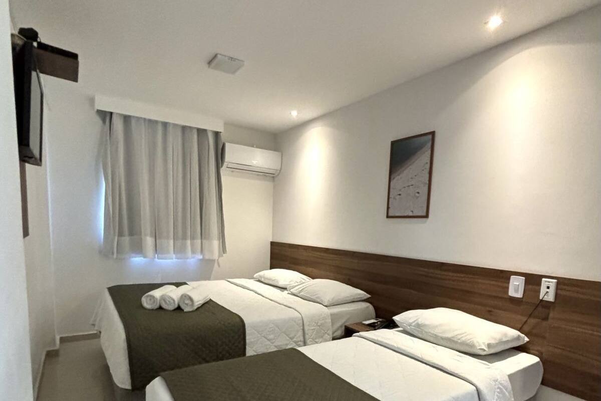 A hotel room with two beds, a window with curtains, an air conditioning unit, and a framed picture on the wall.