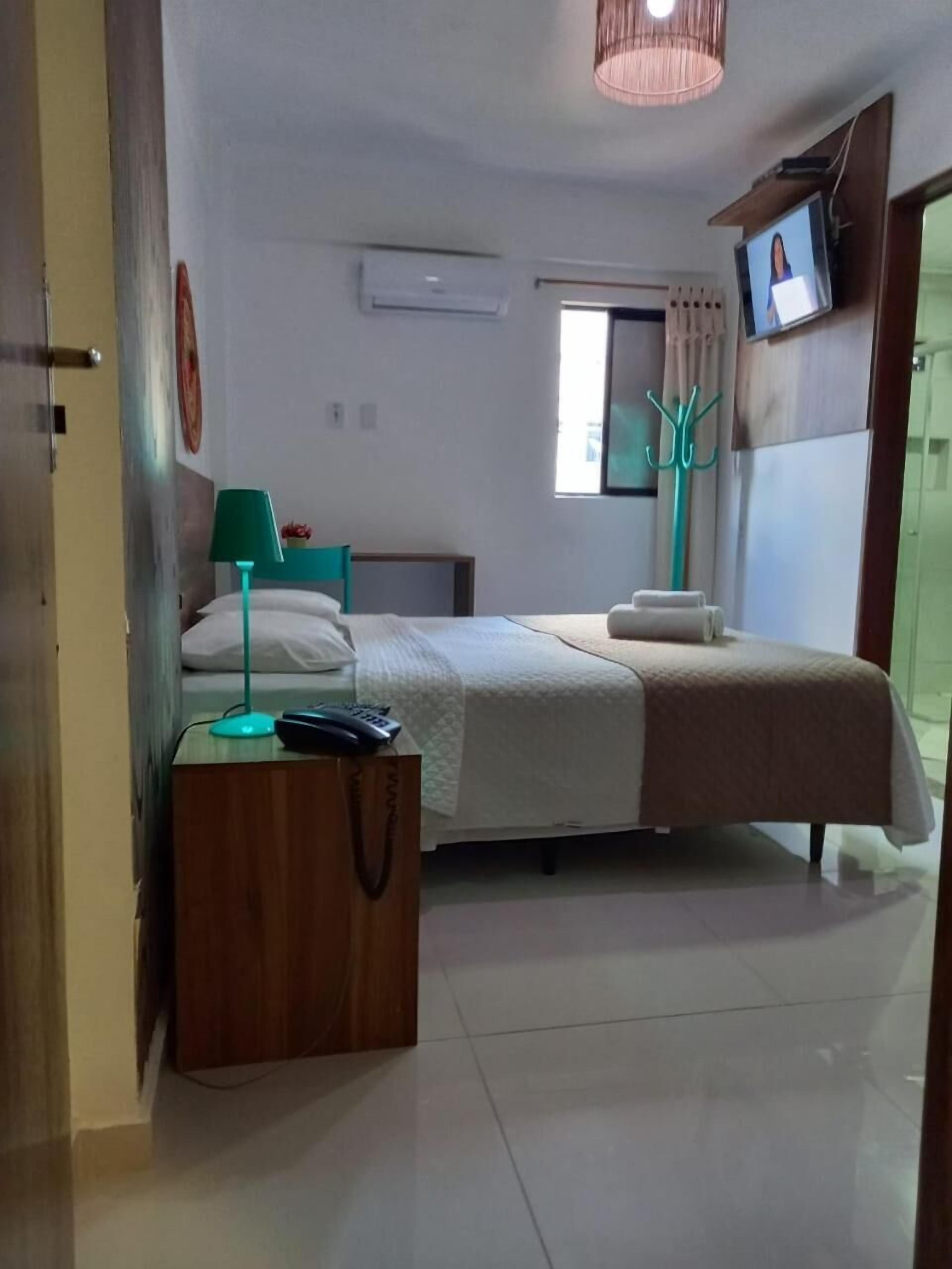 A bedroom with a bed, a desk, a chair, a TV, and a window.
