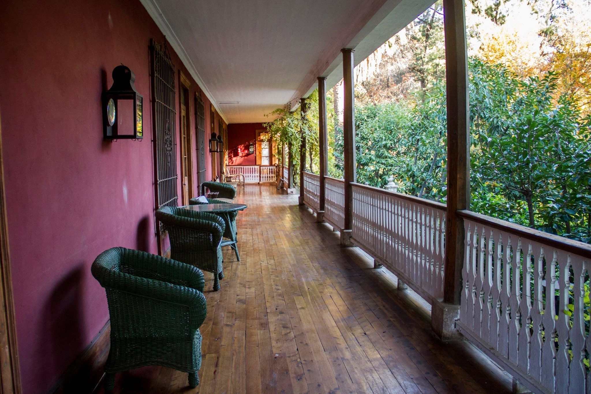 A covered porch with wicker chairs and tables, wooden flooring, and a railing overlooking a garden.